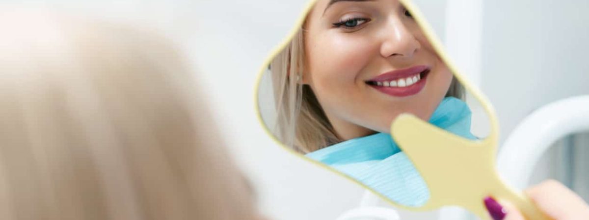 A woman smiles at her reflection in a hand mirror, admiring her beautiful, healthy teeth.