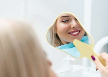 A woman smiles at her reflection in a hand mirror, admiring her beautiful, healthy teeth.