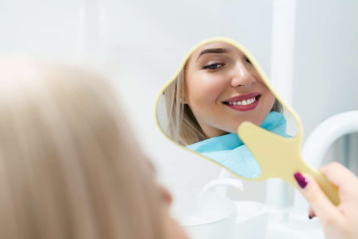 A woman smiles at her reflection in a hand mirror, admiring her beautiful, healthy teeth.