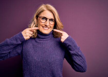 A middle-aged woman dressed for cold weather points at her own beautiful, healthy teeth, enjoying her smile makeover.