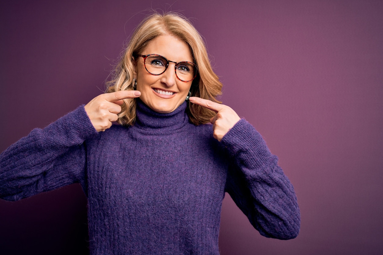 A middle-aged woman dressed for cold weather points at her own beautiful, healthy teeth, enjoying her smile makeover.