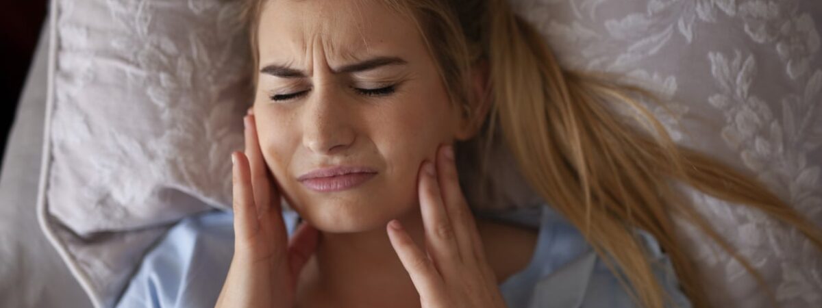 A woman with blonde hair and a blue shirt is in bed, grasping her jaw and experiencing TMJ pain.